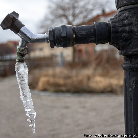 Wasserhahn im Freien, an dem sich ein Eiszapfen gebildet hat, vor einer winterlichen Landschaft im Hintergrund.