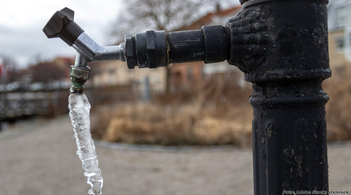 Wasserhahn im Freien, an dem sich ein Eiszapfen gebildet hat, vor einer winterlichen Landschaft im Hintergrund.